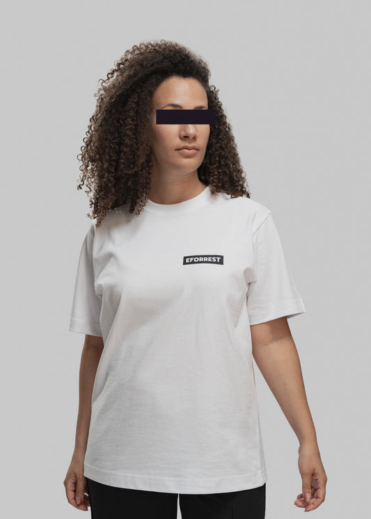 Eine Frau mit lockigem, schulterlangem Haar trägt ein schlichtes weißes T-Shirt mit einem kleinen schwarzen Logo-Patch auf der Brust. Darauf steht in weißen Großbuchstaben „EFORREST“. Die Frau steht frontal zur Kamera, ihr Oberkörper ist leicht nach rechts gedreht, der Blick geht nach links aus dem Bild. Der Hintergrund ist einfarbig hellgrau und neutral gehalten. Ihre Augenpartie ist aus Datenschutzgründen geschwärzt. Die Passform des T-Shirts ist locker, die Ärmel reichen bis knapp über den Ellbogen.