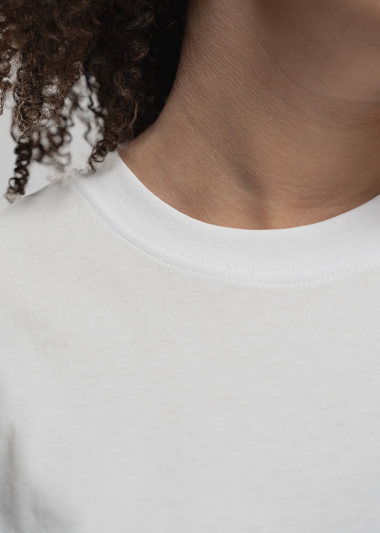 Detailaufnahme des oberen T-Shirt-Bereichs einer Frau mit dunklen, lockigen Haaren. Der Fokus liegt auf dem runden, fein gerippten Kragen des weißen T-Shirts. Der Halsausschnitt ist glatt verarbeitet, das Material wirkt weich und hochwertig. Ein Teil der Hals- und Schulterpartie ist sichtbar, ebenso eine Locke, die leicht auf dem Shirt liegt.