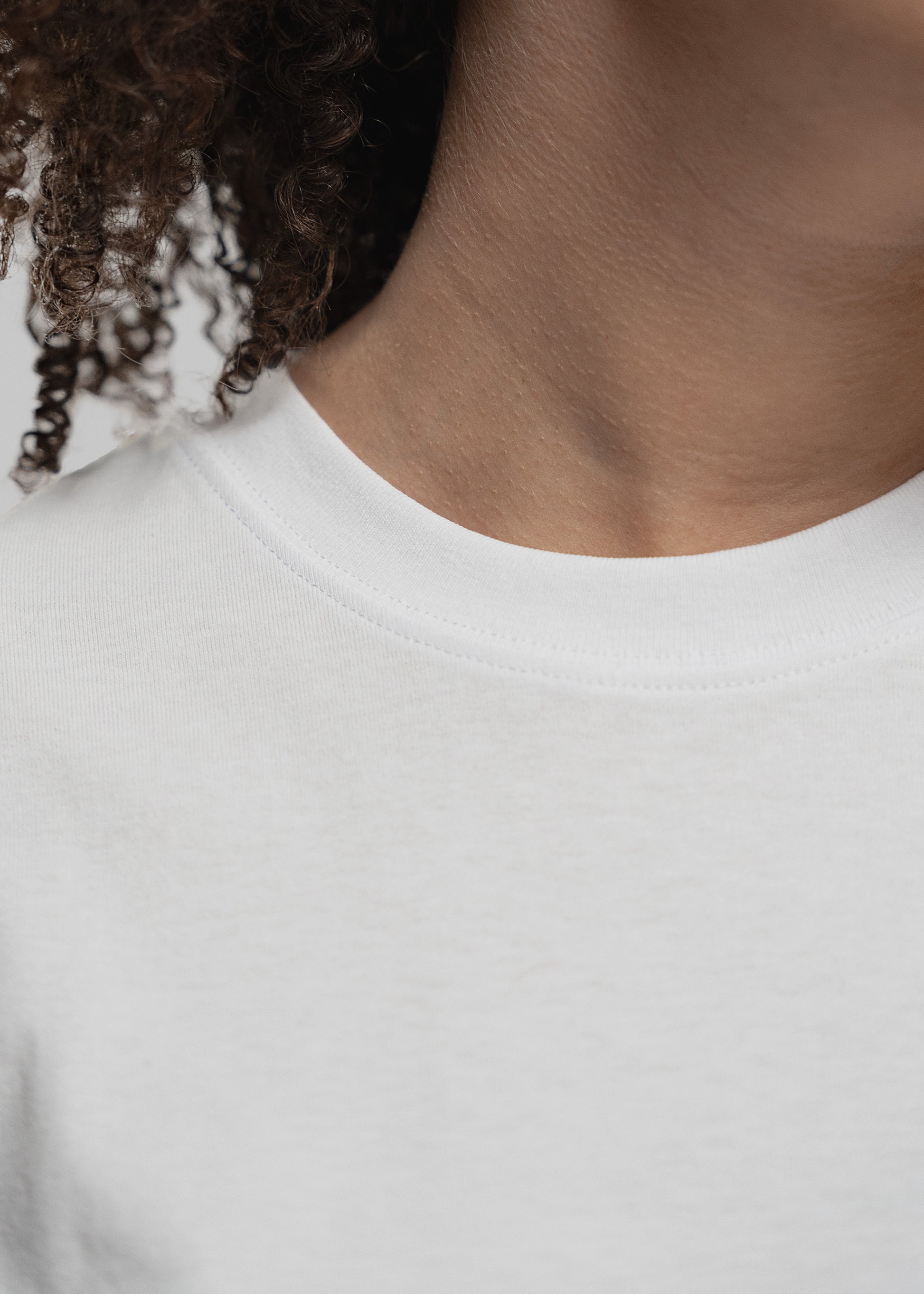 Detailaufnahme des oberen T-Shirt-Bereichs einer Frau mit dunklen, lockigen Haaren. Der Fokus liegt auf dem runden, fein gerippten Kragen des weißen T-Shirts. Der Halsausschnitt ist glatt verarbeitet, das Material wirkt weich und hochwertig. Ein Teil der Hals- und Schulterpartie ist sichtbar, ebenso eine Locke, die leicht auf dem Shirt liegt.