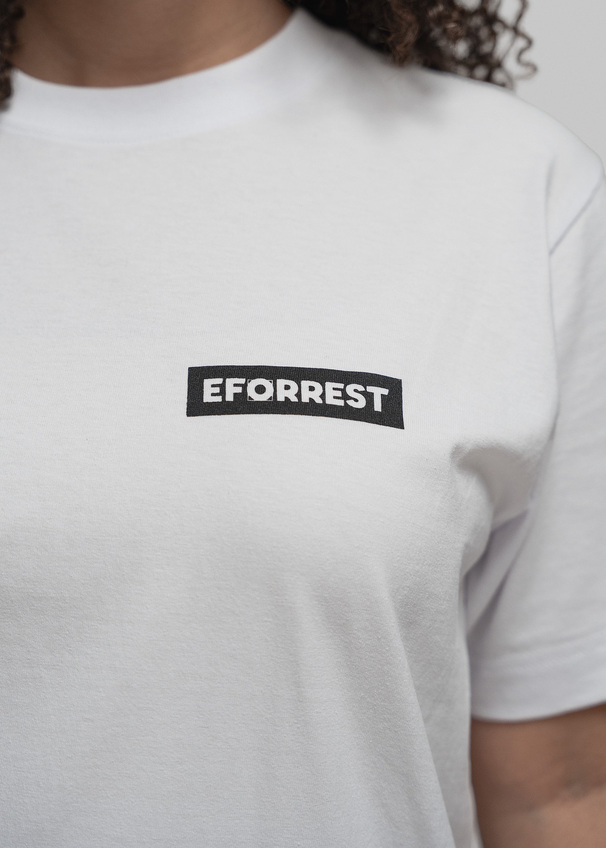 Nahaufnahme eines weißen T-Shirts im Brustbereich. Im Fokus steht ein schwarzes, rechteckiges Logo mit weißer Schrift. In der Mitte des Schriftzugs „EFORREST“ ist das zweite „O“ durch ein stilisiertes Design ersetzt, das an ein Blütenblatt oder einen abstrakten Kreis erinnert. Die Textur des Stoffes sowie die Qualität des Drucks sind deutlich sichtbar. Die Person trägt das Shirt körpernah, mit leichtem Faltenwurf unterhalb der Brust.