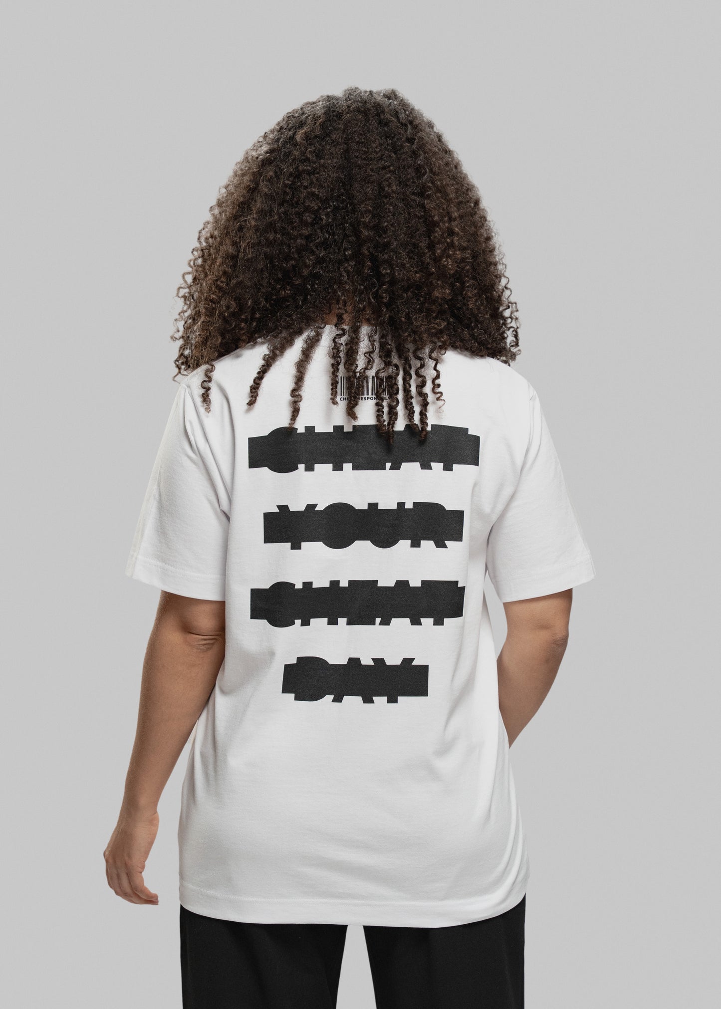 Die abgebildete Person steht mit dem Rücken zur Kamera und trägt ein weißes Oversized-T-Shirt. Der schwarze Aufdruck auf dem Rücken besteht aus vier versetzt gesetzten Textzeilen in grober Blockschrift: „CHEAT YOUR CHEAT DAY“. Über dem Text befindet sich ein minimalistischer Barcode mit kleiner Aufschrift. Die Person hat dunkelbraune, schulterlange Locken und trägt eine schwarze Hose. Der Hintergrund ist neutral hellgrau.