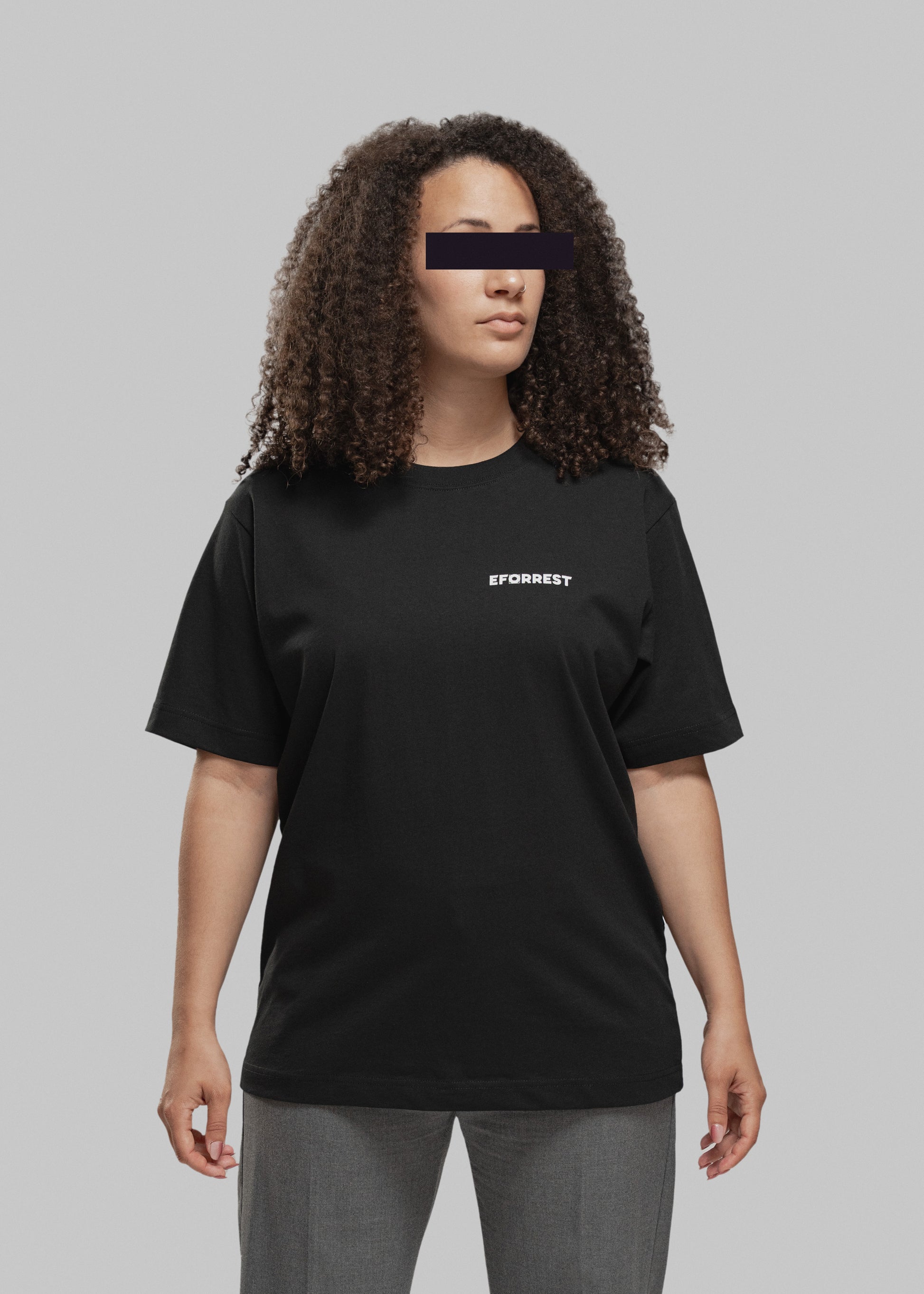 Eine Frau mit mittellanger, voluminös gelockter Frisur steht frontal zur Kamera. Sie trägt ein schwarzes T-Shirt mit weißem, minimalistischen Print auf der linken Brustseite. Dort steht in Großbuchstaben das Wort „EFORREST“, wobei das „O“ durch ein stilisiertes Objekt ersetzt wurde, das wie ein Kameraobjektiv oder technisches Symbol wirkt. Die Frau blickt leicht nach rechts oben. Der Hintergrund ist neutral grau, was den Fokus klar auf die Person und das T-Shirt lenkt.