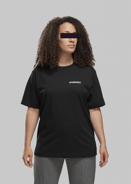 Eine Frau mit mittellanger, voluminös gelockter Frisur steht frontal zur Kamera. Sie trägt ein schwarzes T-Shirt mit weißem, minimalistischen Print auf der linken Brustseite. Dort steht in Großbuchstaben das Wort „EFORREST“, wobei das „O“ durch ein stilisiertes Objekt ersetzt wurde, das wie ein Kameraobjektiv oder technisches Symbol wirkt. Die Frau blickt leicht nach rechts oben. Der Hintergrund ist neutral grau, was den Fokus klar auf die Person und das T-Shirt lenkt.
