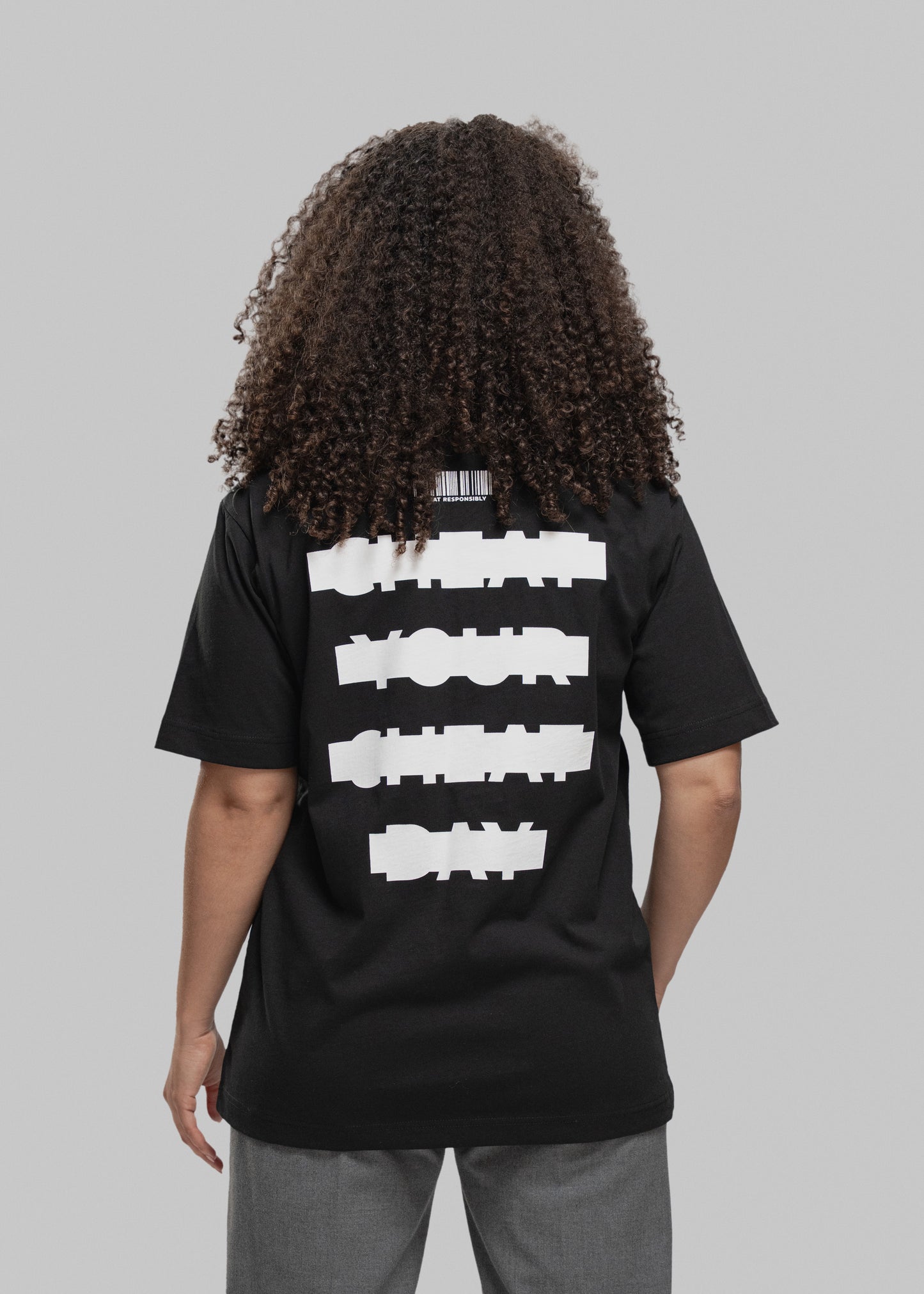 Eine Person mit lockigem, braunem Haar wird von hinten gezeigt. Sie trägt ein schwarzes T-Shirt mit großem weißen Print auf dem Rücken. In vier dicken, horizontal versetzten Balken steht der Slogan: „CHEAT YOUR CHEAT DAY“ – die Wörter sind in Blockschrift geschrieben und wirken bewusst „verpixelt“ bzw. grob maskiert. Über dem Schriftzug befindet sich ein stilisierter Barcode mit dem Text „CHEAT RESPONSIBLY“ darunter. Die Person trägt eine graue Hose, steht vor einem neutral grauen Hintergrund und hält die A