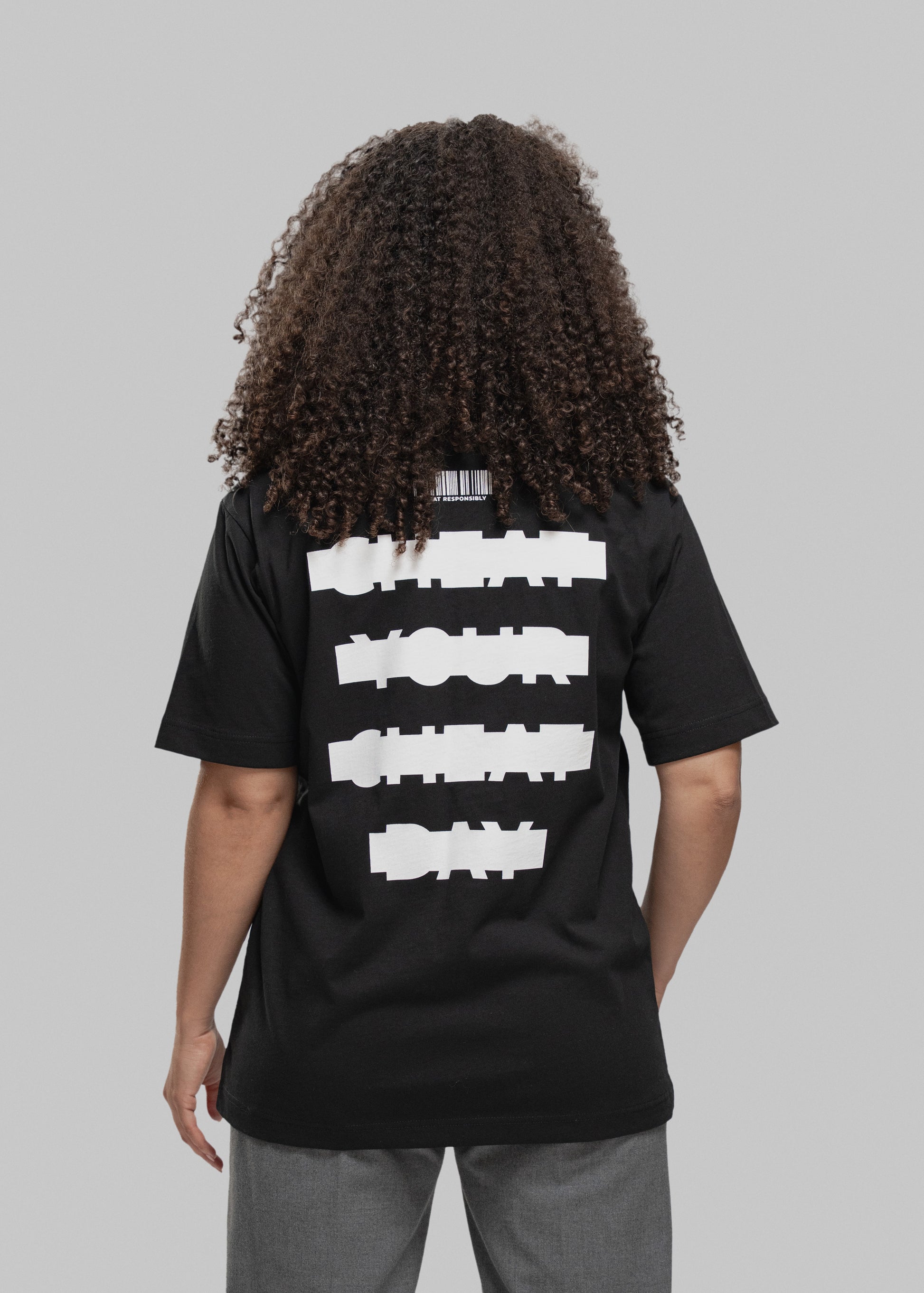 Eine Person mit lockigem, braunem Haar wird von hinten gezeigt. Sie trägt ein schwarzes T-Shirt mit großem weißen Print auf dem Rücken. In vier dicken, horizontal versetzten Balken steht der Slogan: „CHEAT YOUR CHEAT DAY“ – die Wörter sind in Blockschrift geschrieben und wirken bewusst „verpixelt“ bzw. grob maskiert. Über dem Schriftzug befindet sich ein stilisierter Barcode mit dem Text „CHEAT RESPONSIBLY“ darunter. Die Person trägt eine graue Hose, steht vor einem neutral grauen Hintergrund und hält die A