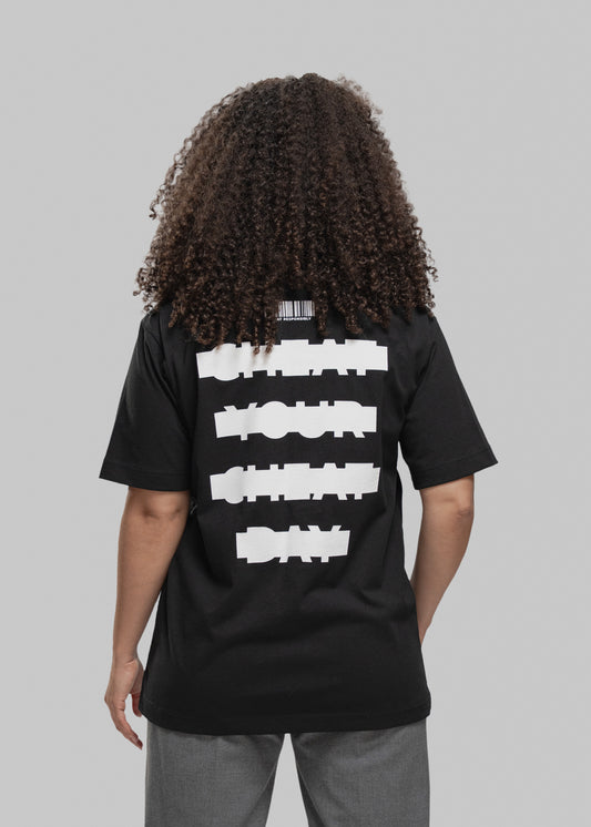 Eine Person mit lockigem, braunem Haar wird von hinten gezeigt. Sie trägt ein schwarzes T-Shirt mit großem weißen Print auf dem Rücken. In vier dicken, horizontal versetzten Balken steht der Slogan: „CHEAT YOUR CHEAT DAY“ – die Wörter sind in Blockschrift geschrieben und wirken bewusst „verpixelt“ bzw. grob maskiert. Über dem Schriftzug befindet sich ein stilisierter Barcode mit dem Text „CHEAT RESPONSIBLY“ darunter. Die Person trägt eine graue Hose, steht vor einem neutral grauen Hintergrund und hält die A