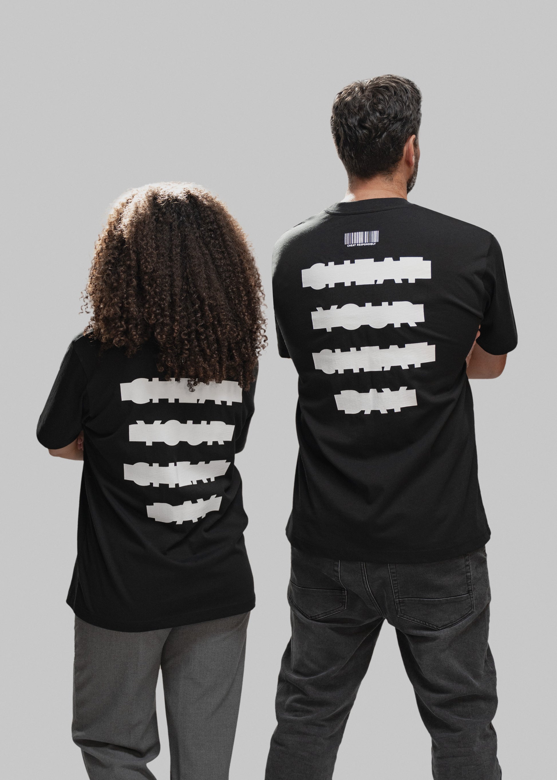 Zwei Personen stehen Rücken an Rücken vor einem neutral grauen Hintergrund. Beide tragen schwarze T-Shirts mit identischem weißem Print auf dem Rücken: Vier große, horizontale Schriftbalken zeigen die Worte „CHEAT YOUR CHEAT DAY“ in verzerrter, blockartiger Schrift. Über dem Text befindet sich ein kleiner Barcode mit der Aufschrift „CHEAT RESPONSIBLY“. Links steht eine Person mit lockigem, schulterlangem Haar und grauer Hose, rechts ein Mann mit kurzen Haaren und dunkler Jeans. Die Pose beider Personen wirk