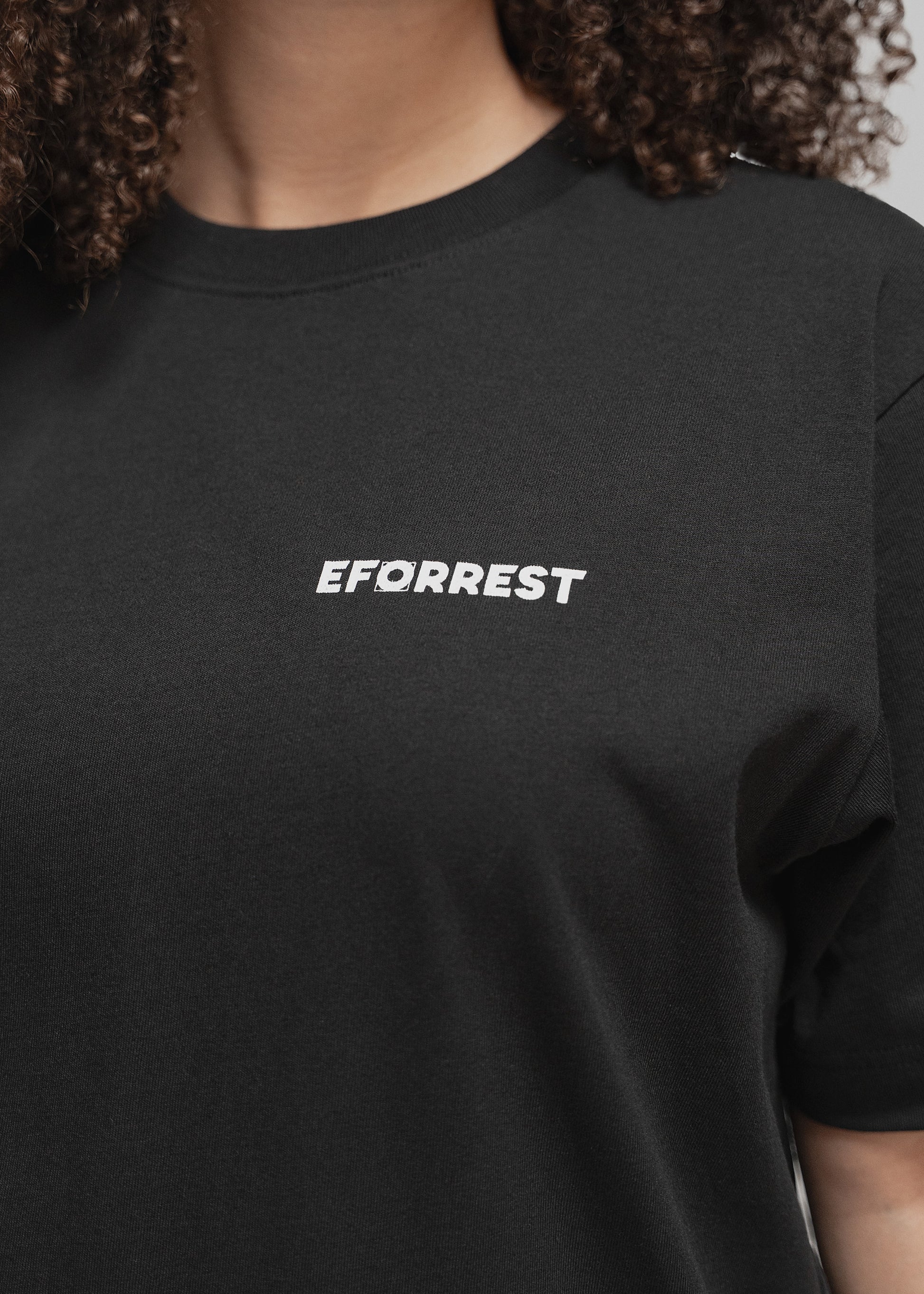 Nahaufnahme des oberen Torsos einer Person mit lockigem Haar, die ein schwarzes T-Shirt trägt. Auf der linken Brustseite befindet sich der weiße Schriftzug „EFORREST“. Der Buchstabe „O“ ist als stilisiertes Zahnrad oder Kreissymbol mit grafischer Struktur gestaltet. Der Ausschnitt des T-Shirts ist rund und sitzt nah am Hals. Der Hintergrund ist neutral grau, sodass der Fokus vollständig auf dem T-Shirt und dem Logo liegt.