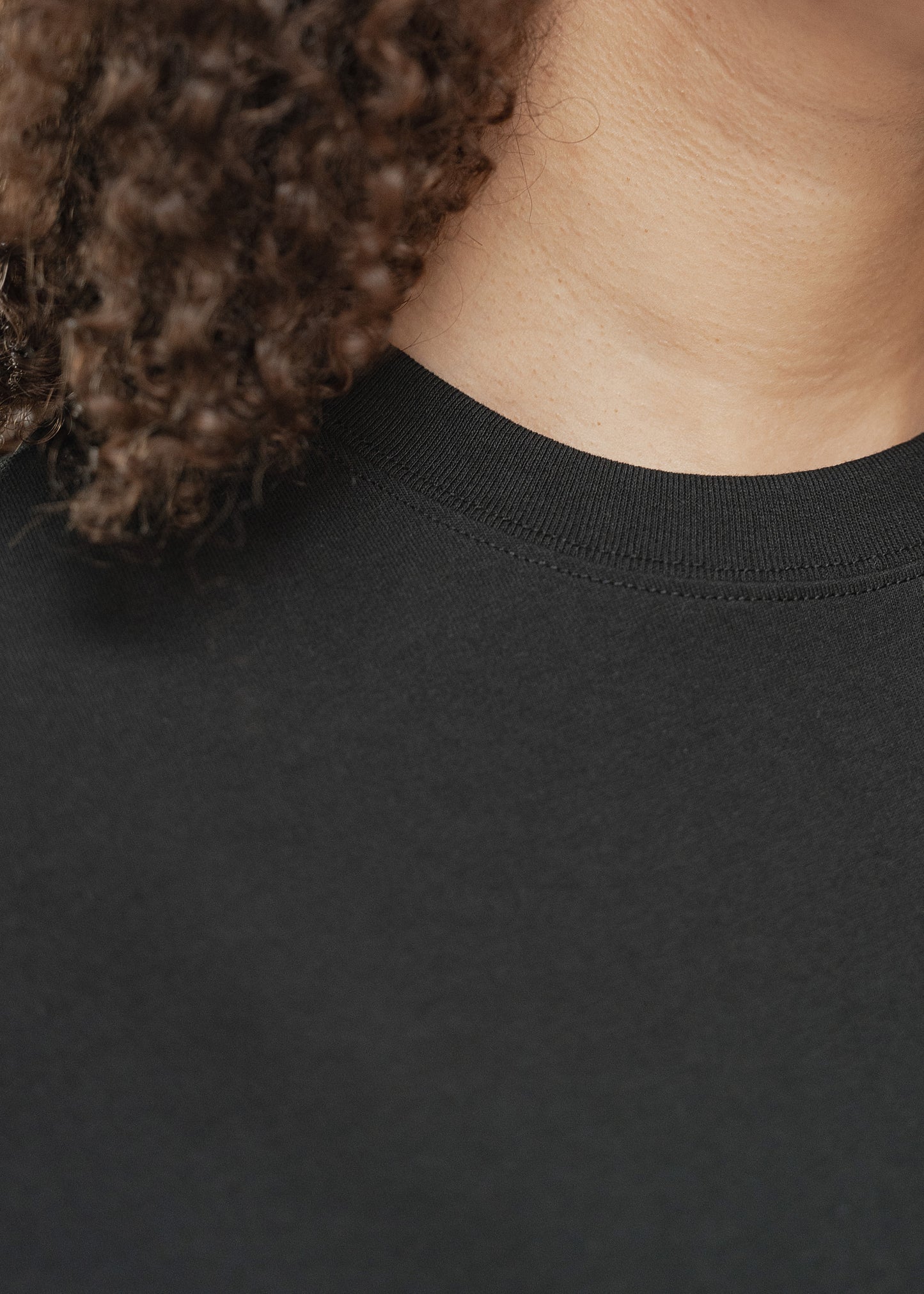 Nahaufnahme des Halsausschnitts eines schwarzen T-Shirts, getragen von einer Person mit lockigem Haar. Zu sehen ist der Rundhalsausschnitt mit feiner Doppelnaht sowie die feine Textur des Baumwollstoffs. Links im Bild hängen einige Locken herab, rechts ist ein Ausschnitt des Halses und der unteren Gesichtspartie sichtbar. Der Hintergrund ist neutral und unscharf, um das Material und die Passform des Kragens in den Vordergrund zu stellen.