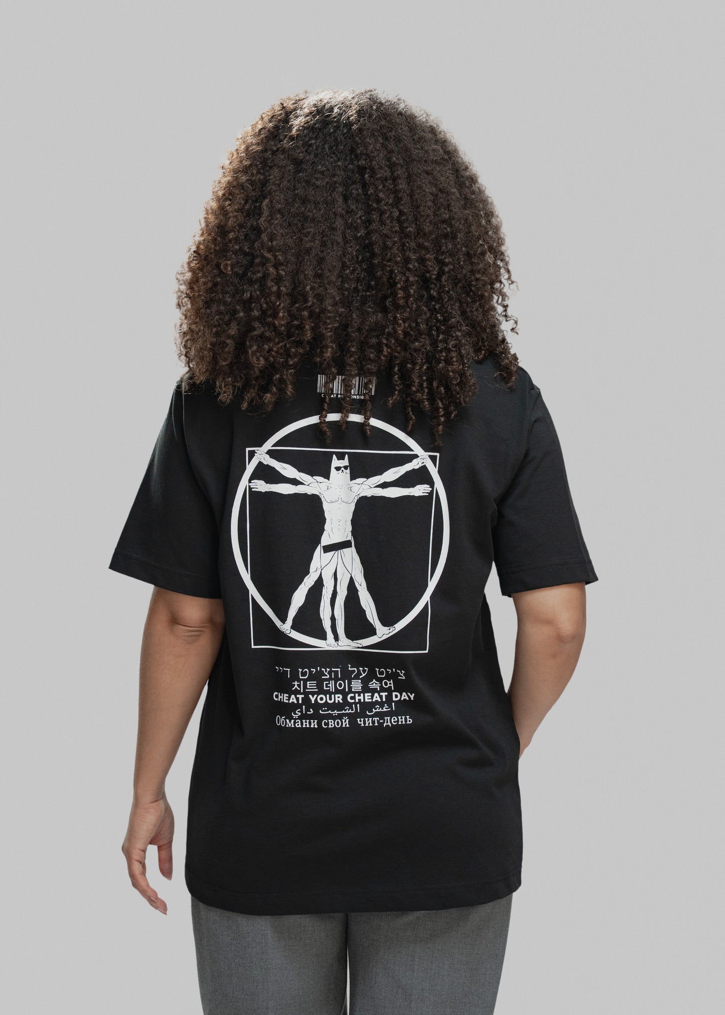 Eine Person mit lockigem Haar steht mit dem Rücken zur Kamera und trägt ein schwarzes T-Shirt. Auf dem Rücken ist ein auffälliges weißes Motiv zu sehen: Eine muskulöse Comicfigur mit Maske – stark an den „Vitruvianischen Menschen“ von Leonardo da Vinci angelehnt – steht mit ausgestreckten Armen und Beinen in einem Kreis und Quadrat. Der Charakter trägt nur eine enge Shorts. Oberhalb des Motivs befindet sich ein stilisierter Barcode mit der Aufschrift „CHEAT RESPONSIBLY“. Darunter steht der Satz „CHEAT YOUR 