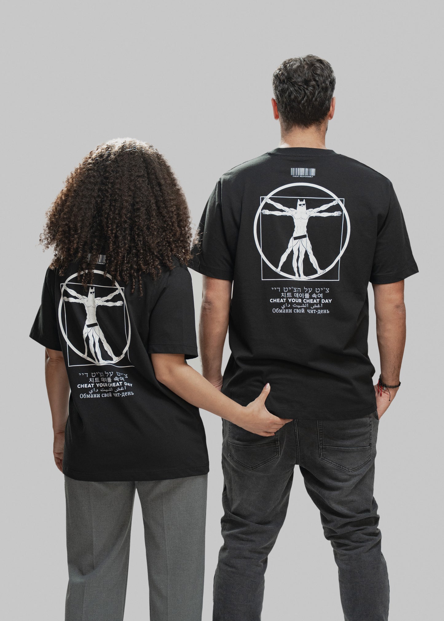 Zwei Personen stehen nebeneinander mit dem Rücken zur Kamera. Beide tragen ein schwarzes T-Shirt mit demselben auffälligen Rückendruck: Im Zentrum ist eine vitruvianisch dargestellte Superhelden-Figur zu sehen, die stark an den „Vitruvianischen Menschen“ erinnert – allerdings mit Maske, Sixpack und Comic-Anmutung. Darüber befindet sich ein weißer Barcode mit der Aufschrift „CHEAT RESPONSIBLY“. Unterhalb des Motivs steht der Spruch „CHEAT YOUR CHEAT DAY“ in sechs verschiedenen Sprachen (u. a. Hebräisch, Kore
