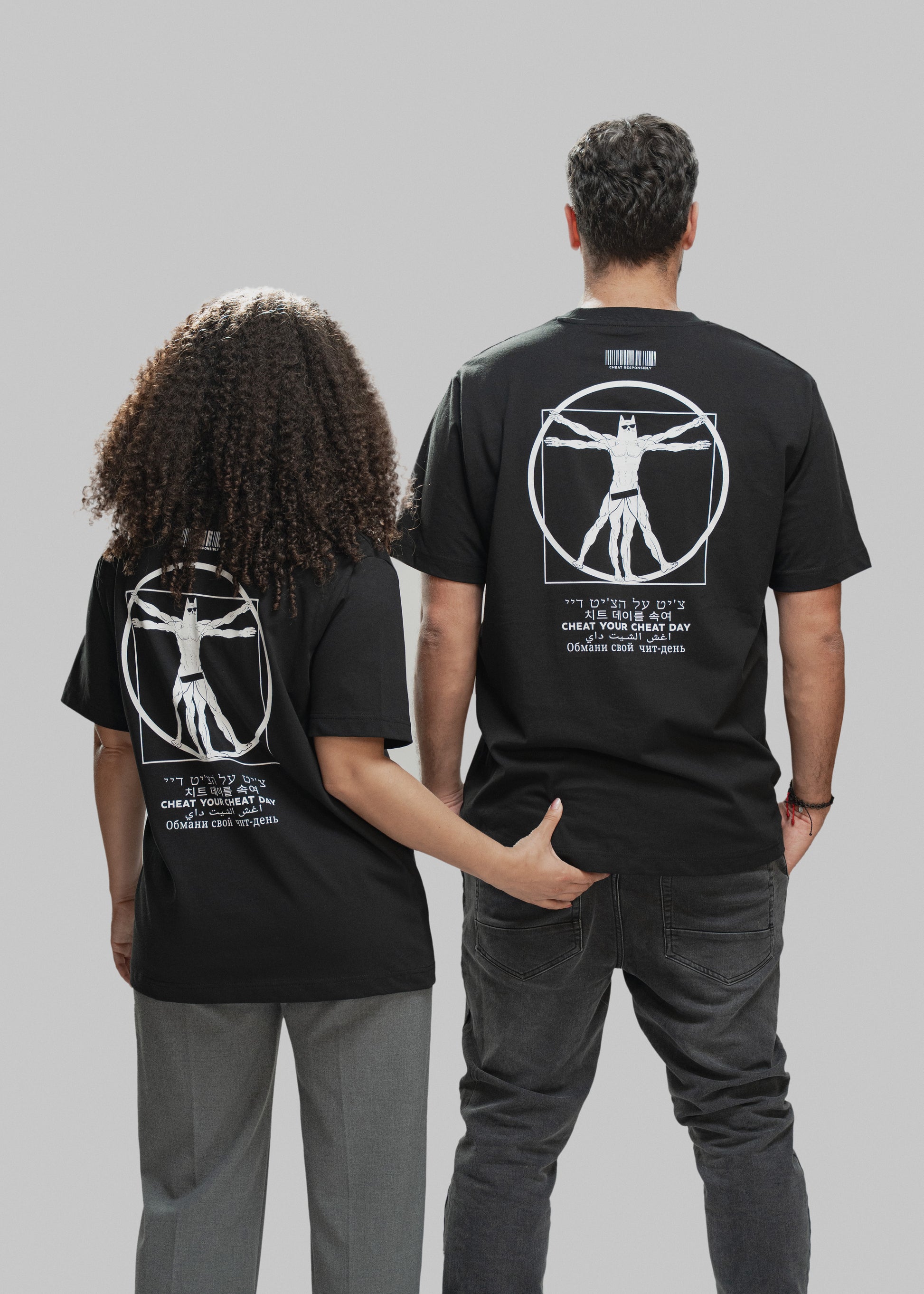 Zwei Personen stehen nebeneinander mit dem Rücken zur Kamera. Beide tragen ein schwarzes T-Shirt mit demselben auffälligen Rückendruck: Im Zentrum ist eine vitruvianisch dargestellte Superhelden-Figur zu sehen, die stark an den „Vitruvianischen Menschen“ erinnert – allerdings mit Maske, Sixpack und Comic-Anmutung. Darüber befindet sich ein weißer Barcode mit der Aufschrift „CHEAT RESPONSIBLY“. Unterhalb des Motivs steht der Spruch „CHEAT YOUR CHEAT DAY“ in sechs verschiedenen Sprachen (u. a. Hebräisch, Kore