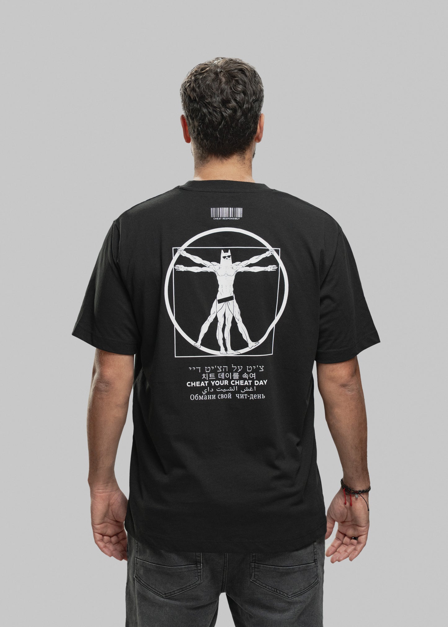 Ein Mann steht mit dem Rücken zur Kamera und trägt ein schwarzes T-Shirt mit einem auffälligen weißen Print auf der Rückseite. Im Zentrum des Designs befindet sich eine muskulöse Figur im Vitruvianischen-Stil – eine Mischung aus Da Vincis berühmtem „Vitruvianischen Menschen“ und einer Superhelden-Figur mit Maske, Sixpack und breiten Schultern. Die Figur steht in einem Kreis und Quadrat, beide Elemente sind weiß umrandet. Oberhalb der Grafik ist ein Barcode mit dem Schriftzug „CHEAT RESPONSIBLY“. Unterhalb d