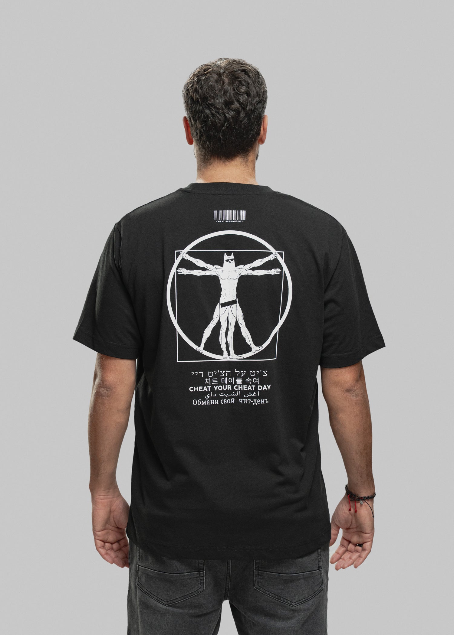 Ein Mann steht mit dem Rücken zur Kamera und trägt ein schwarzes T-Shirt mit einem auffälligen weißen Print auf der Rückseite. Im Zentrum des Designs befindet sich eine muskulöse Figur im Vitruvianischen-Stil – eine Mischung aus Da Vincis berühmtem „Vitruvianischen Menschen“ und einer Superhelden-Figur mit Maske, Sixpack und breiten Schultern. Die Figur steht in einem Kreis und Quadrat, beide Elemente sind weiß umrandet. Oberhalb der Grafik ist ein Barcode mit dem Schriftzug „CHEAT RESPONSIBLY“. Unterhalb d