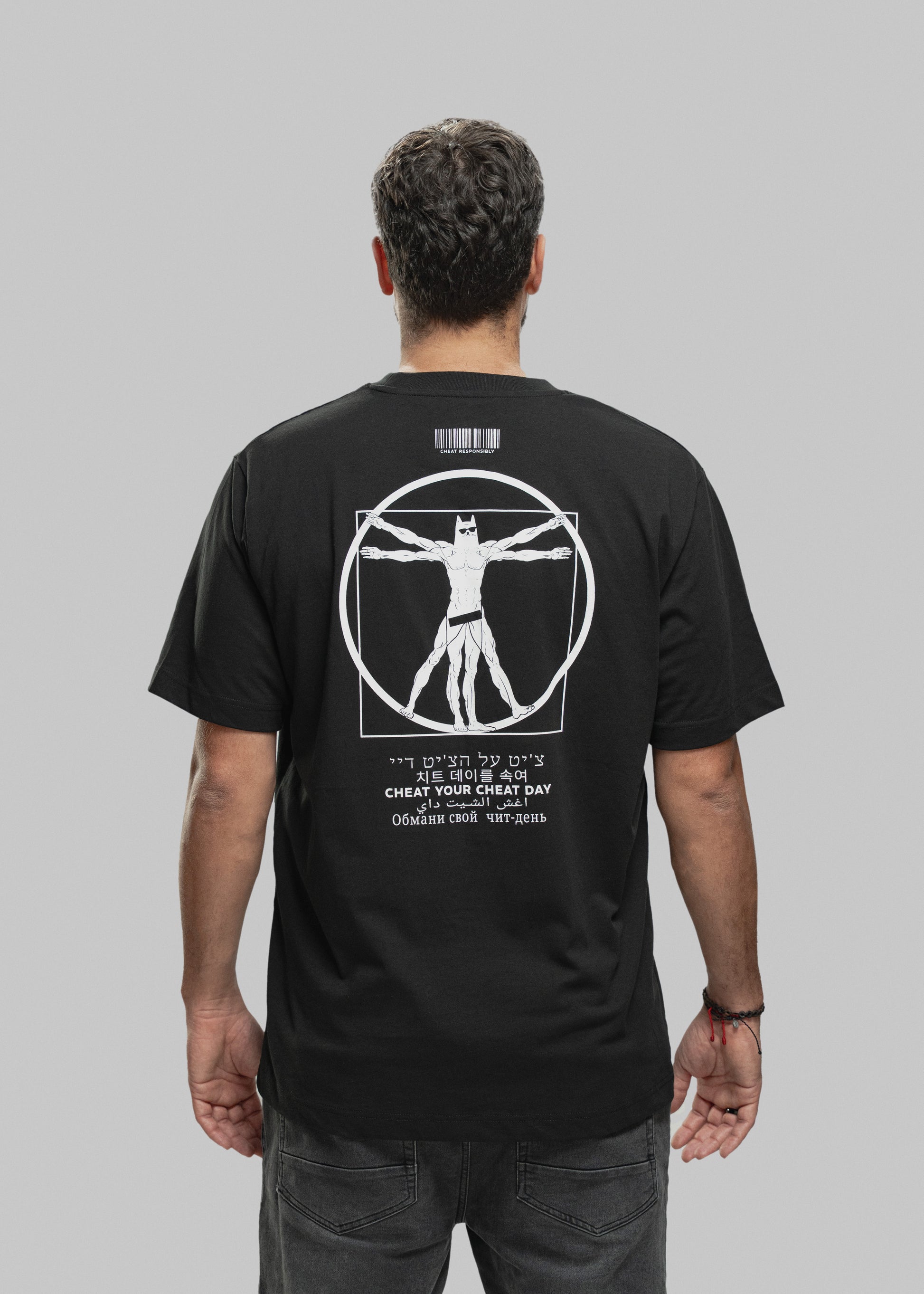 Ein Mann steht mit dem Rücken zur Kamera und trägt ein schwarzes T-Shirt mit einem auffälligen weißen Print auf der Rückseite. Im Zentrum des Designs befindet sich eine muskulöse Figur im Vitruvianischen-Stil – eine Mischung aus Da Vincis berühmtem „Vitruvianischen Menschen“ und einer Superhelden-Figur mit Maske, Sixpack und breiten Schultern. Die Figur steht in einem Kreis und Quadrat, beide Elemente sind weiß umrandet. Oberhalb der Grafik ist ein Barcode mit dem Schriftzug „CHEAT RESPONSIBLY“. Unterhalb d