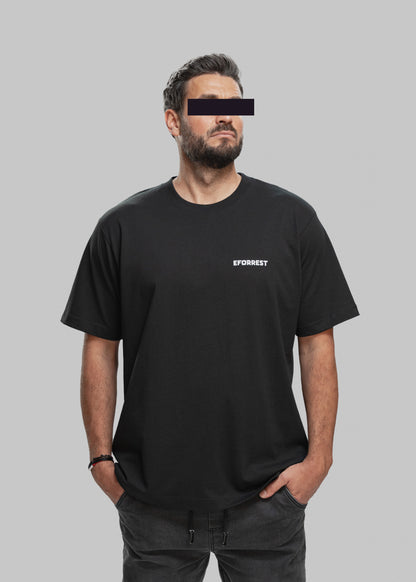 Ein Mann mit dunklem, kurzem Haar, gepflegtem Bart und leicht gebräunter Haut steht frontal vor einem hellgrauen Hintergrund. Er trägt ein schlichtes, schwarzes T-Shirt mit einem weißen „EFORREST“-Logo auf der linken Brustseite. Seine linke Hand steckt in der Hosentasche, die rechte hängt locker herunter. Er blickt leicht nach rechts oben, sein Gesichtsausdruck wirkt konzentriert und ruhig. Die Augenpartie ist durch einen schwarzen Balken anonymisiert. Das Outfit wird durch eine dunkle Jogginghose mit Korde