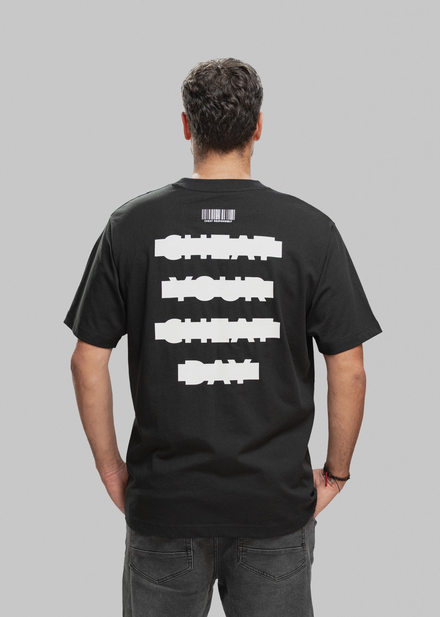 Ein Mann mit kurzen, dunkelbraunen Haaren wird von hinten gezeigt. Er trägt ein schwarzes T-Shirt mit einem auffälligen weißen Statement-Print auf dem Rücken. In vier horizontal angeordneten Balken steht in versetzter, blockartiger Schrift: „CHEAT YOUR CHEAT DAY“. Über dem Text ist ein stilisierter Barcode abgebildet, darunter in kleiner Schrift der Slogan „CHEAT RESPONSIBLY“. Der Mann trägt dunkle Jeans, hat die Hände in den Hosentaschen und steht vor neutral grauem Studiohintergrund.