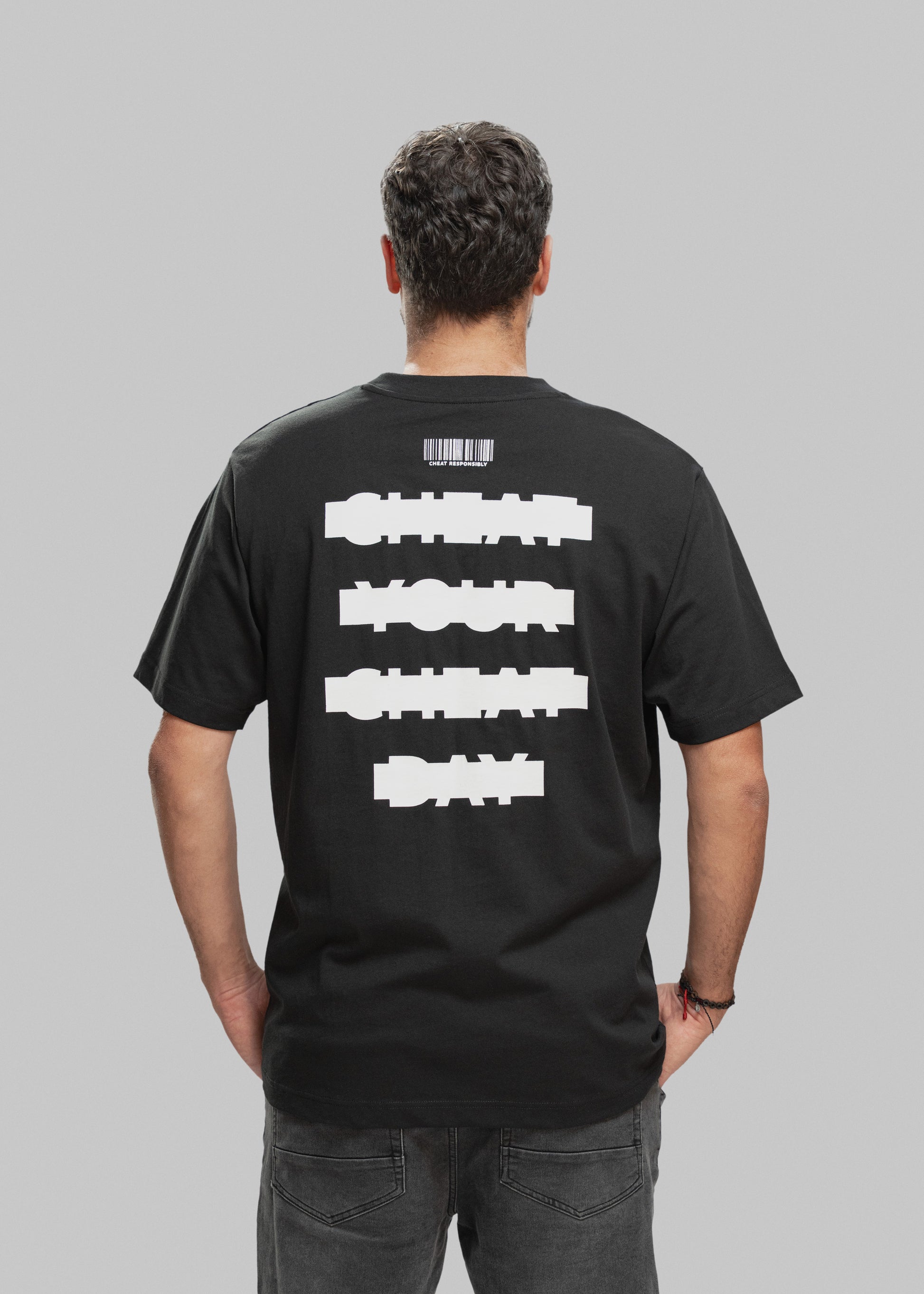 Ein Mann mit kurzen, dunkelbraunen Haaren wird von hinten gezeigt. Er trägt ein schwarzes T-Shirt mit einem auffälligen weißen Statement-Print auf dem Rücken. In vier horizontal angeordneten Balken steht in versetzter, blockartiger Schrift: „CHEAT YOUR CHEAT DAY“. Über dem Text ist ein stilisierter Barcode abgebildet, darunter in kleiner Schrift der Slogan „CHEAT RESPONSIBLY“. Der Mann trägt dunkle Jeans, hat die Hände in den Hosentaschen und steht vor neutral grauem Studiohintergrund.