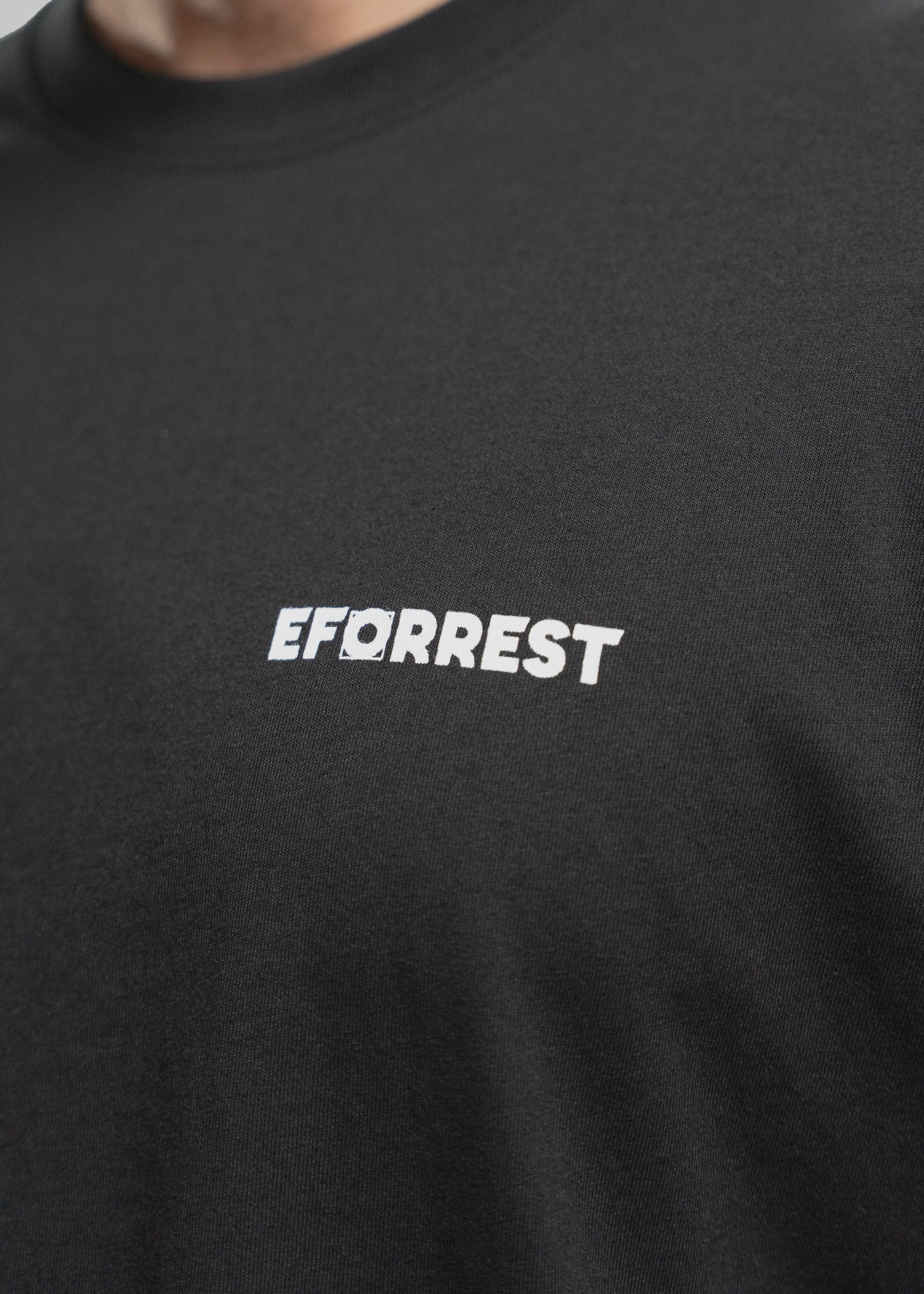 Nahaufnahme eines schwarzen T-Shirts, das mittig die weiße Aufschrift „EFORREST“ zeigt. Der Fokus liegt auf dem Logo, bei dem der Buchstabe „O“ durch eine stilisierte Linse oder Zieloptik ersetzt ist – ein Designelement, das visuell an Fotografie erinnert. Der Schriftzug hebt sich klar vom schwarzen Stoff ab. Der obere Bereich des Bildes zeigt einen Ausschnitt vom Hals und Kinn einer Person mit hellem Teint und Bartstoppeln. Die Textur des T-Shirts ist fein und leicht elastisch erkennbar.