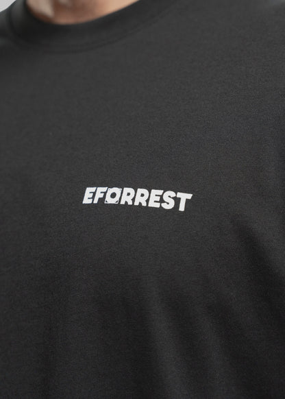 Nahaufnahme eines schwarzen T-Shirts, das mittig die weiße Aufschrift „EFORREST“ zeigt. Der Fokus liegt auf dem Logo, bei dem der Buchstabe „O“ durch eine stilisierte Linse oder Zieloptik ersetzt ist – ein Designelement, das visuell an Fotografie erinnert. Der Schriftzug hebt sich klar vom schwarzen Stoff ab. Der obere Bereich des Bildes zeigt einen Ausschnitt vom Hals und Kinn einer Person mit hellem Teint und Bartstoppeln. Die Textur des T-Shirts ist fein und leicht elastisch erkennbar.