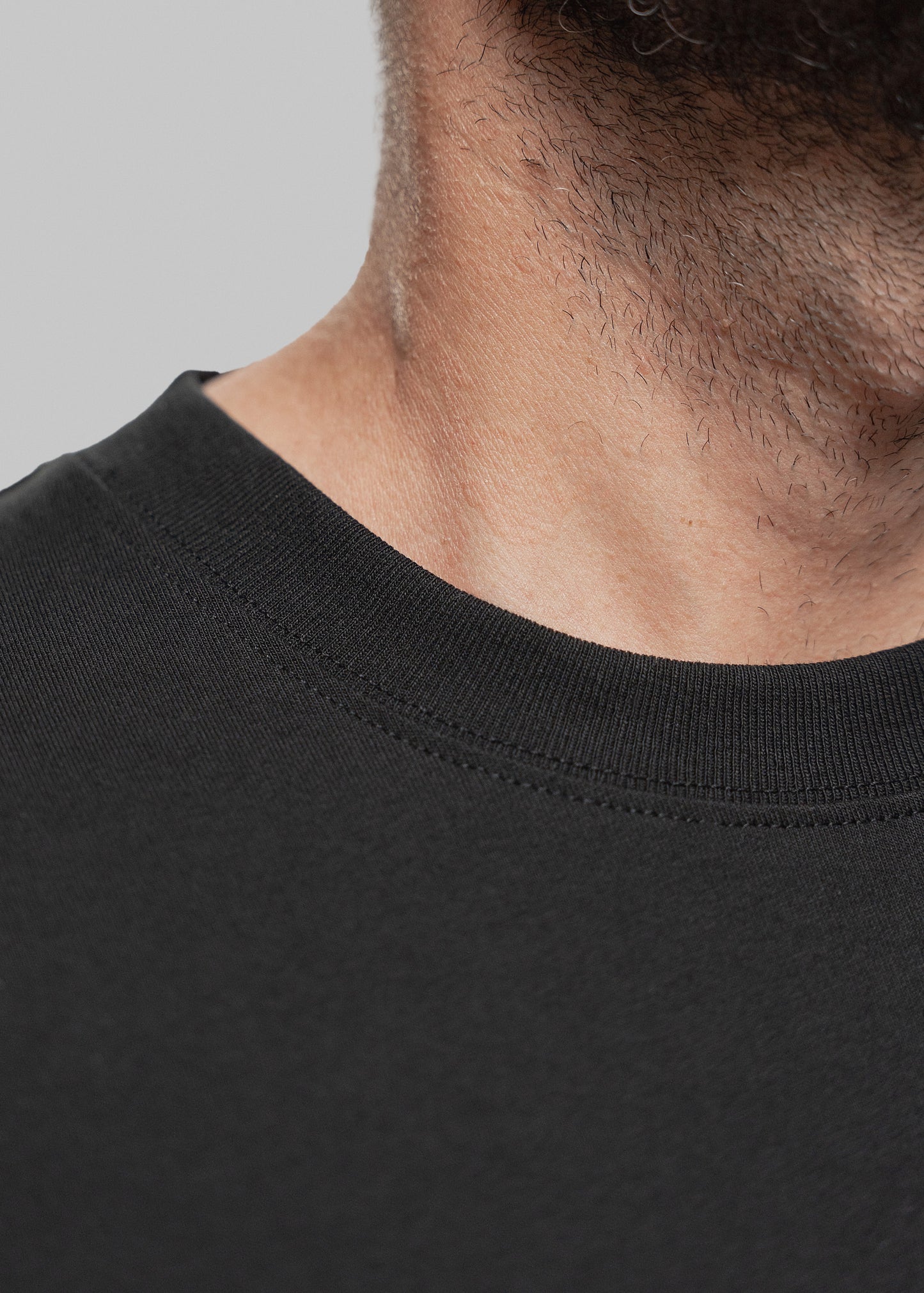 Detailaufnahme eines schwarzen T-Shirts mit Fokus auf den Halsbereich eines Mannes mit kurzem Bartwuchs. Sichtbar ist der fein gerippte Rundhalsausschnitt des Shirts mit doppelter Naht. Die Textur des Stoffes ist glatt, matt und engmaschig, was auf hochwertige Verarbeitung hinweist. Der Teint des Mannes ist hell bis leicht gebräunt. Die Perspektive zeigt ausschließlich den unteren Teil seines Bartes, Halses und Schlüsselbeins – der Bildausschnitt bleibt auf Stoffqualität und Tragekomfort fokussiert.