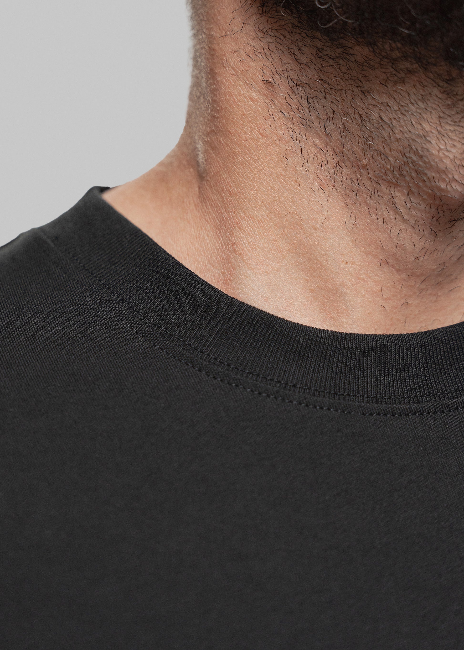 Detailaufnahme eines schwarzen T-Shirts mit Fokus auf den Halsbereich eines Mannes mit kurzem Bartwuchs. Sichtbar ist der fein gerippte Rundhalsausschnitt des Shirts mit doppelter Naht. Die Textur des Stoffes ist glatt, matt und engmaschig, was auf hochwertige Verarbeitung hinweist. Der Teint des Mannes ist hell bis leicht gebräunt. Die Perspektive zeigt ausschließlich den unteren Teil seines Bartes, Halses und Schlüsselbeins – der Bildausschnitt bleibt auf Stoffqualität und Tragekomfort fokussiert.
