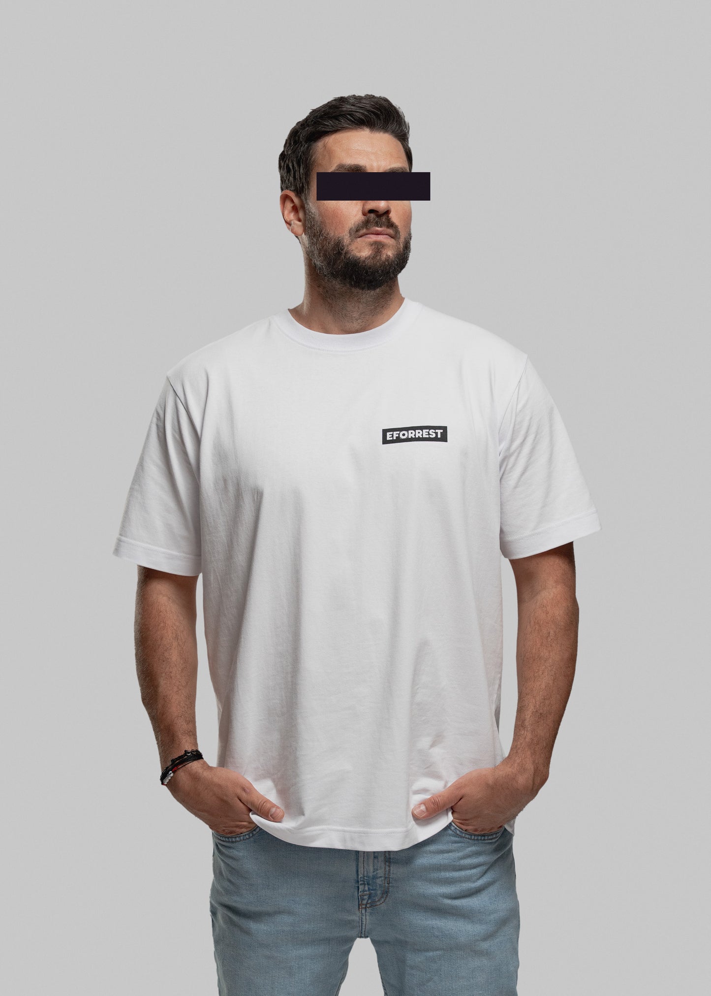 Auf dem Bild steht eine männlich gelesene Person vor neutralgrauem Hintergrund. Sie trägt ein locker geschnittenes weißes T-Shirt mit einem kleinen schwarzen EFORREST-Schriftzug in Großbuchstaben auf der linken Brust. Das Gesicht ist teilweise anonymisiert: Die Augenpartie ist mit einem schwarzen Balken zensiert. Die Person hat beide Hände in den Hosentaschen und trägt eine helle Jeans sowie ein Armband am linken Handgelenk. Der Stil ist modern, schlicht und streetwear-inspiriert.