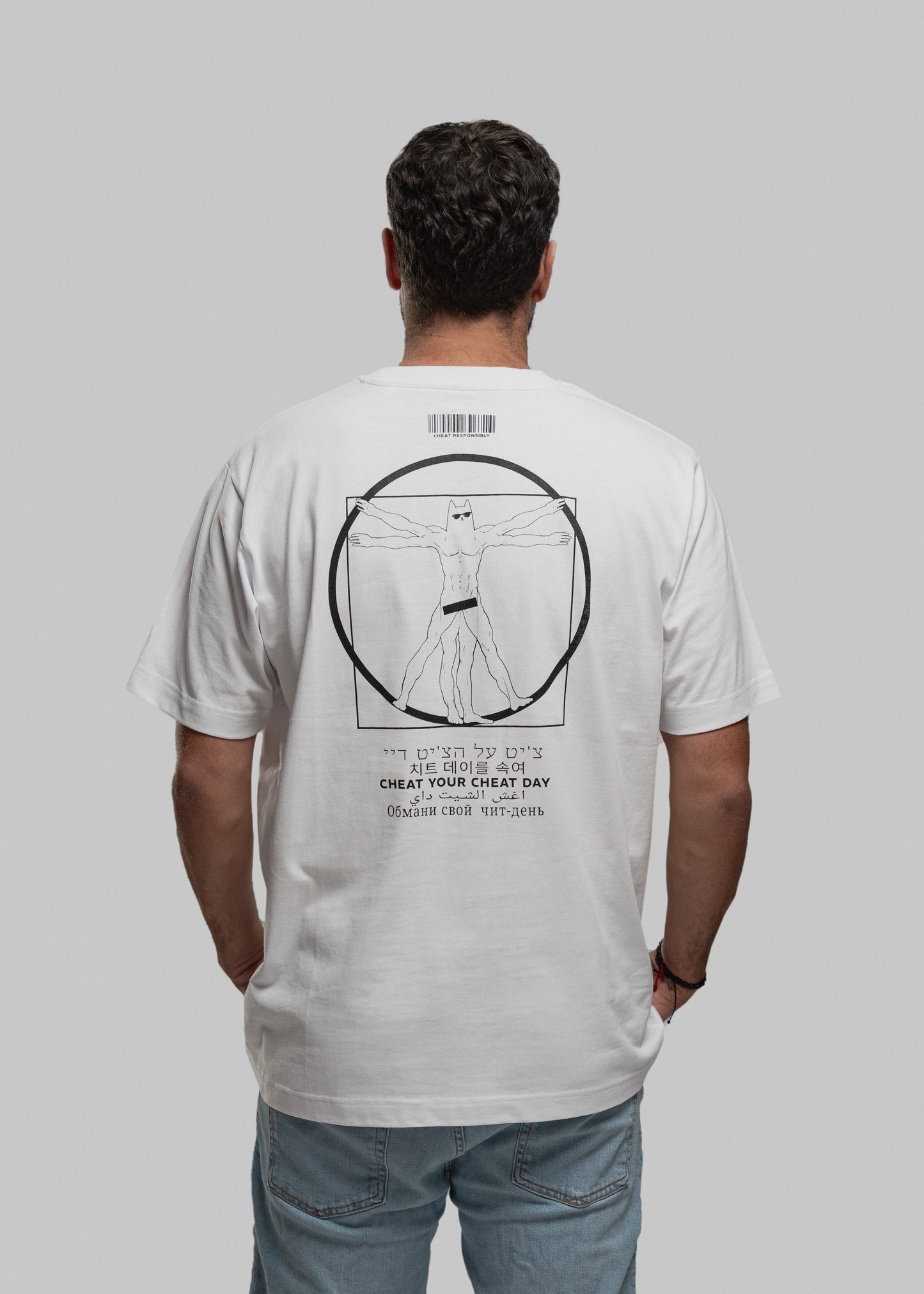 Die fotografierte Person trägt ein weißes T-Shirt mit einem großflächigen, zentralen Aufdruck auf dem Rücken. Zu sehen ist eine humorvolle Version von Leonardos Vitruvianischem Menschen: muskulöser Körper, mehrfach dargestellte Gliedmaßen, Katzenkopf, schwarze Unterhose. Über der Figur ist ein schmaler Barcode mit der Aufschrift „CHEAT RESPONSIBLY.“ zu sehen. Unter der Grafik steht der Marken-Slogan „CHEAT YOUR CHEAT DAY“ in mehreren Sprachen, darunter Hebräisch, Koreanisch, Arabisch, Kyrillisch und Lateinb