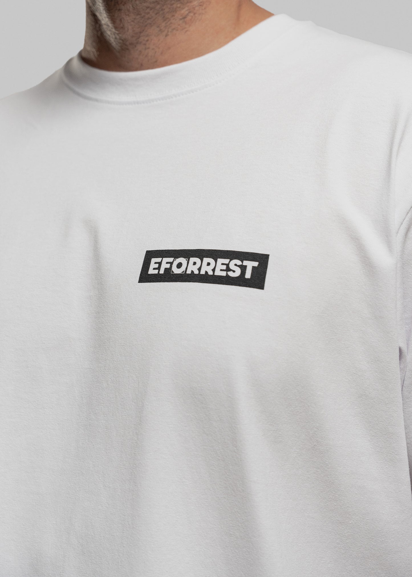 Auf dem Bild ist der obere Brustbereich einer Person zu sehen, die ein schlichtes weißes T-Shirt trägt. Links auf Brusthöhe befindet sich ein kleines schwarzes Rechteck mit dem weißen Schriftzug „EFORREST“. Das „O“ im Schriftzug ist als rundes, grafisches Zeichen gestaltet und hebt sich visuell vom Rest des Textes ab. Der Kragen ist rund und sauber vernäht, der Hintergrund ist neutralgrau.