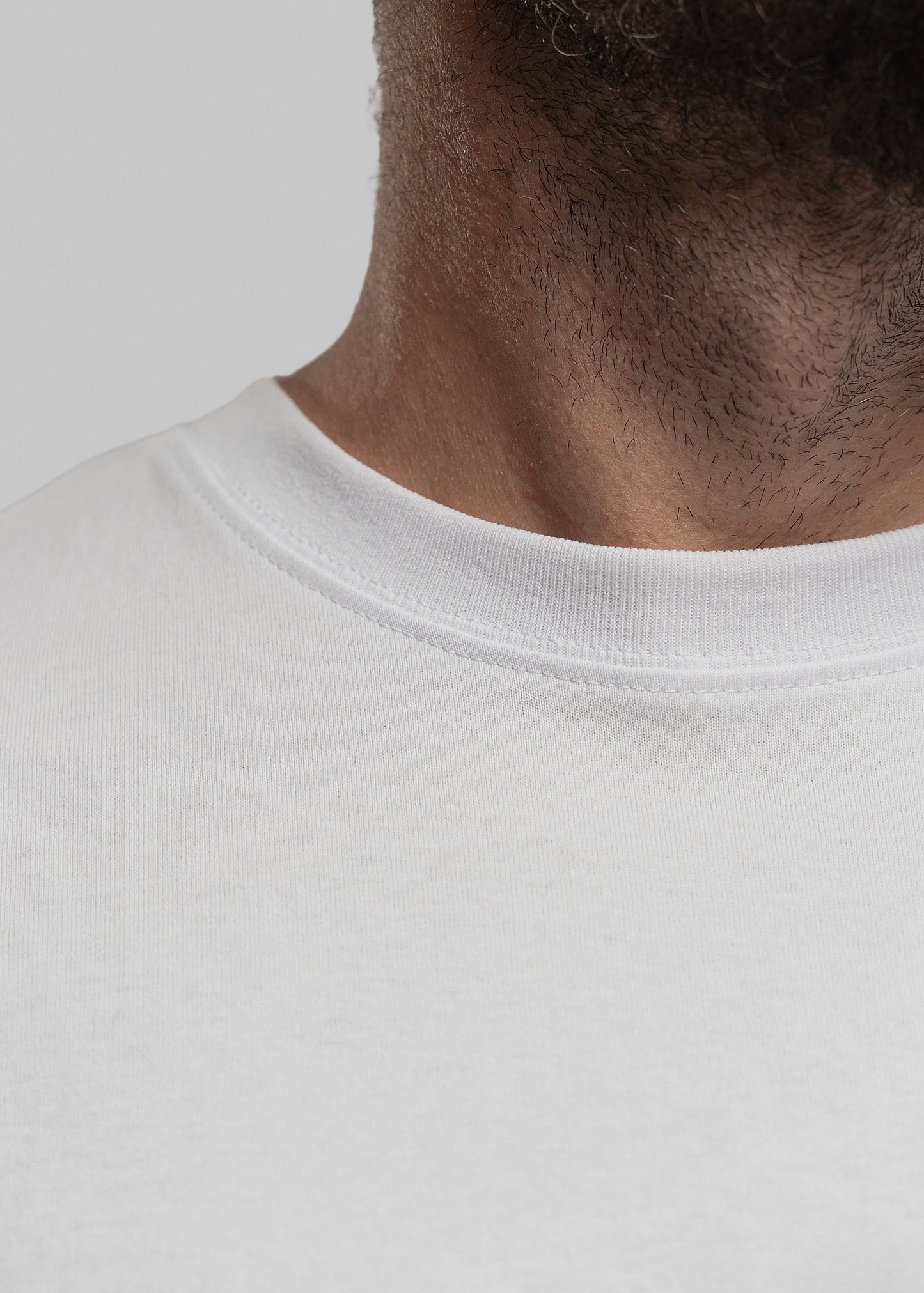 Das Bild zeigt eine Detailaufnahme des Rundhalsausschnitts eines weißen T-Shirts. Im Fokus steht die hochwertige Verarbeitung des gerippten Kragens sowie die feine, saubere Naht. Der Halsausschnitt liegt glatt am Hals des Trägers an. Der Hintergrund ist neutral grau, die Perspektive zeigt den unteren Hals- und oberen Brustbereich des Trägers mit leichtem Bartansatz.