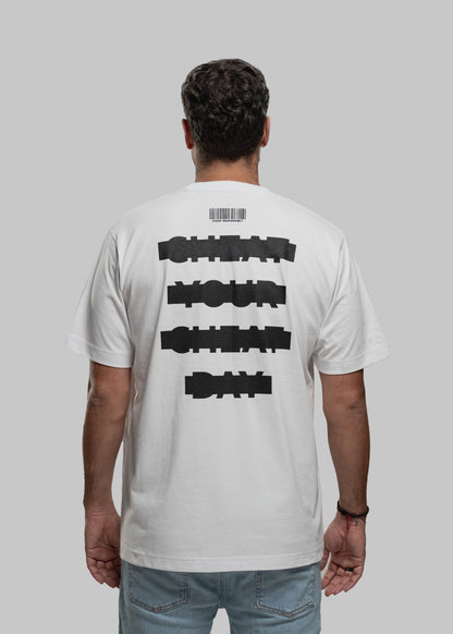 Die abgebildete Person steht mit dem Rücken zur Kamera und trägt ein weißes Oversized-T-Shirt. Der schwarze Aufdruck auf dem Rücken besteht aus vier versetzt gesetzten Textzeilen in grober Blockschrift: „CHEAT YOUR CHEAT DAY“. Über dem Text befindet sich ein minimalistischer Barcode mit kleiner Aufschrift. Die Person hat dunkelbraune, kurze Haare und trägt eine hellblaue Jeans. Der Hintergrund ist neutral hellgrau.