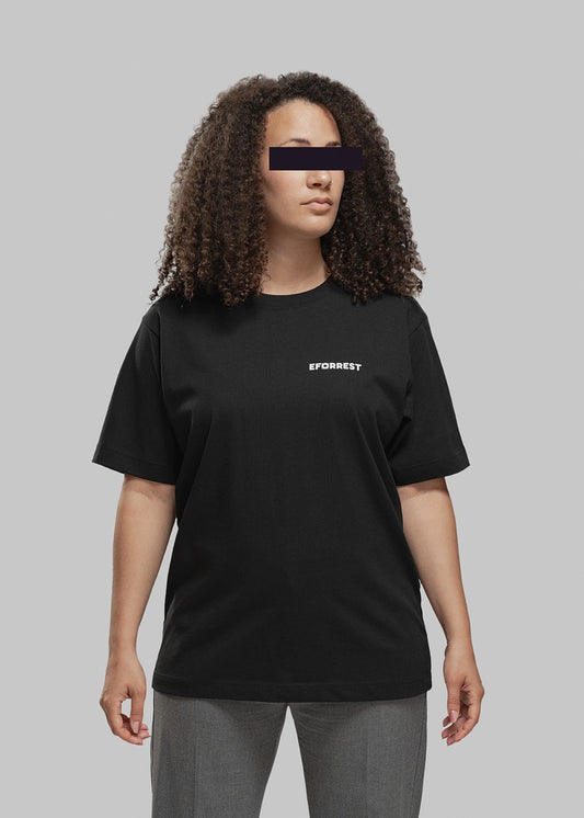 Eine Frau mit mittellanger, voluminös gelockter Frisur steht frontal zur Kamera. Sie trägt ein schwarzes T-Shirt mit weißem, minimalistischen Print auf der linken Brustseite. Dort steht in Großbuchstaben das Wort „EFORREST“, wobei das „O“ durch ein stilisiertes Objekt ersetzt wurde, das wie ein Kameraobjektiv oder technisches Symbol wirkt. Die Frau blickt leicht nach rechts oben. Der Hintergrund ist neutral grau, was den Fokus klar auf die Person und das T-Shirt lenkt.