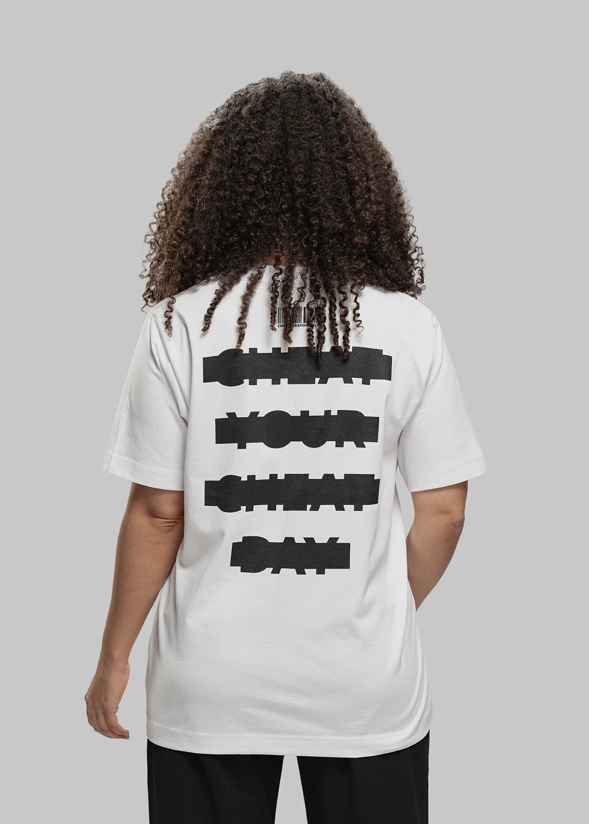 Die abgebildete Person steht mit dem Rücken zur Kamera und trägt ein weißes Oversized-T-Shirt. Der schwarze Aufdruck auf dem Rücken besteht aus vier versetzt gesetzten Textzeilen in grober Blockschrift: „CHEAT YOUR CHEAT DAY“. Über dem Text befindet sich ein minimalistischer Barcode mit kleiner Aufschrift. Die Person hat dunkelbraune, schulterlange Locken und trägt eine schwarze Hose. Der Hintergrund ist neutral hellgrau.