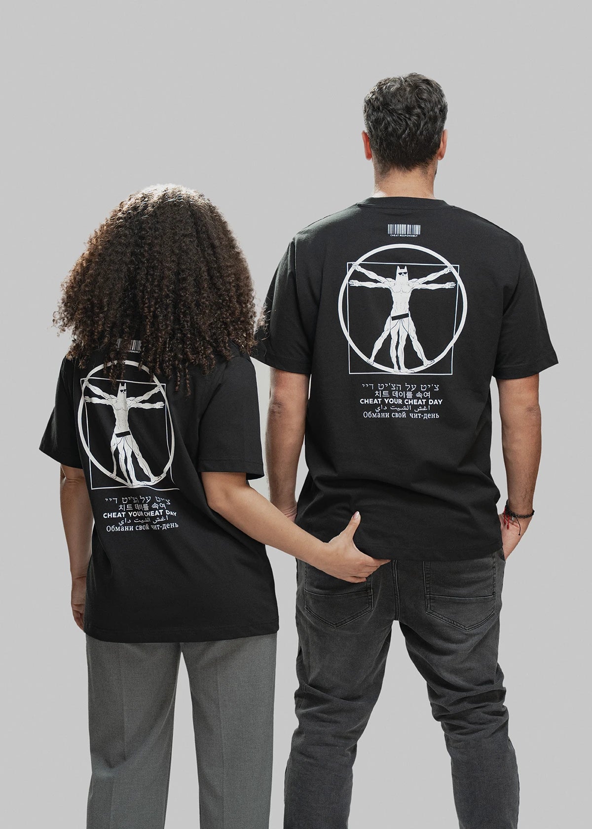 Zwei Personen stehen nebeneinander mit dem Rücken zur Kamera. Beide tragen ein schwarzes T-Shirt mit demselben auffälligen Rückendruck: Im Zentrum ist eine vitruvianisch dargestellte Superhelden-Figur zu sehen, die stark an den „Vitruvianischen Menschen“ erinnert – allerdings mit Maske, Sixpack und Comic-Anmutung. Darüber befindet sich ein weißer Barcode mit der Aufschrift „CHEAT RESPONSIBLY“. Unterhalb des Motivs steht der Spruch „CHEAT YOUR CHEAT DAY“ in sechs verschiedenen Sprachen (u. a. Hebräisch, Kore