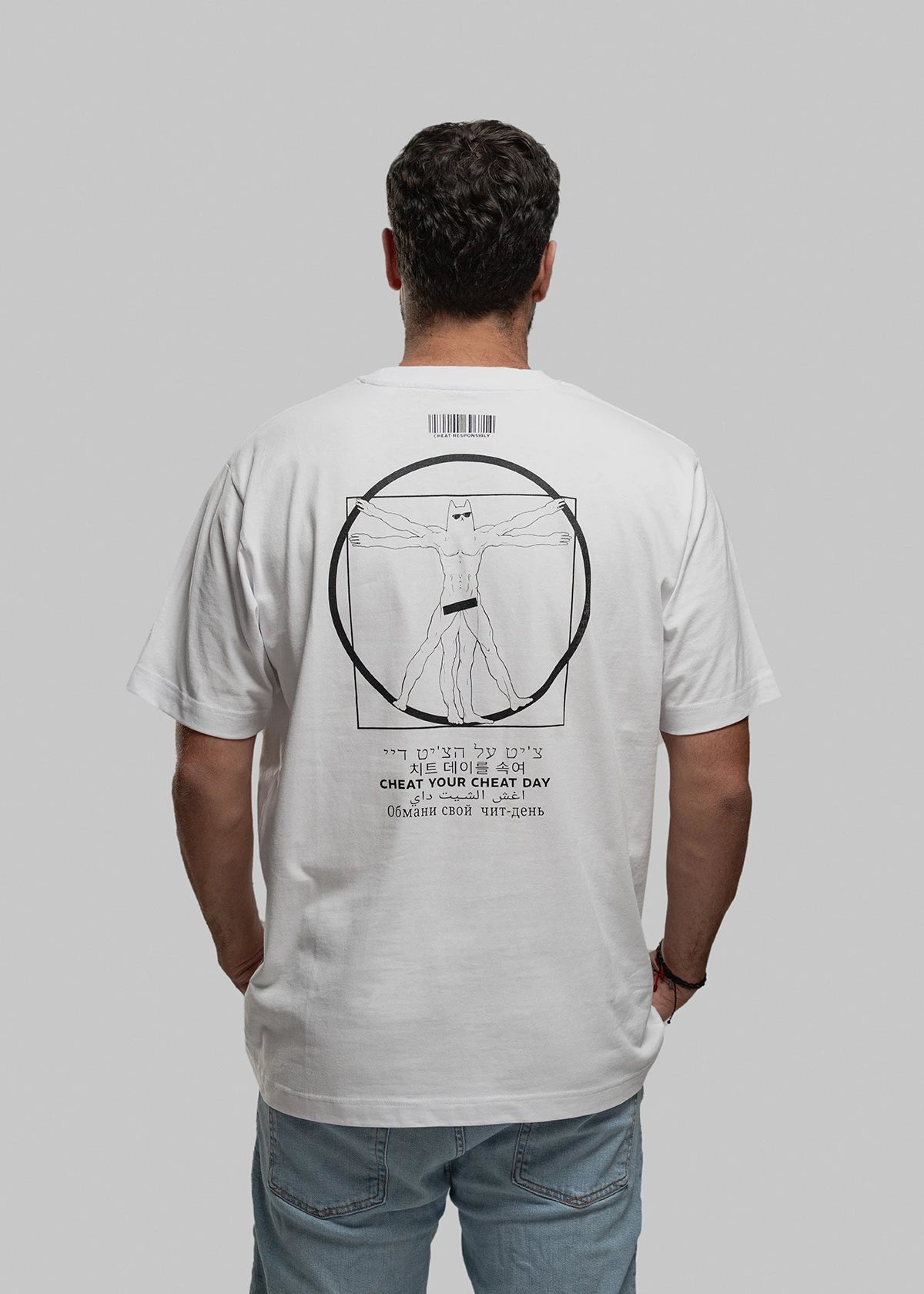 Die fotografierte Person trägt ein weißes T-Shirt mit einem großflächigen, zentralen Aufdruck auf dem Rücken. Zu sehen ist eine humorvolle Version von Leonardos Vitruvianischem Menschen: muskulöser Körper, mehrfach dargestellte Gliedmaßen, Katzenkopf, schwarze Unterhose. Über der Figur ist ein schmaler Barcode mit der Aufschrift „CHEAT RESPONSIBLY.“ zu sehen. Unter der Grafik steht der Marken-Slogan „CHEAT YOUR CHEAT DAY“ in mehreren Sprachen, darunter Hebräisch, Koreanisch, Arabisch, Kyrillisch und Lateinb